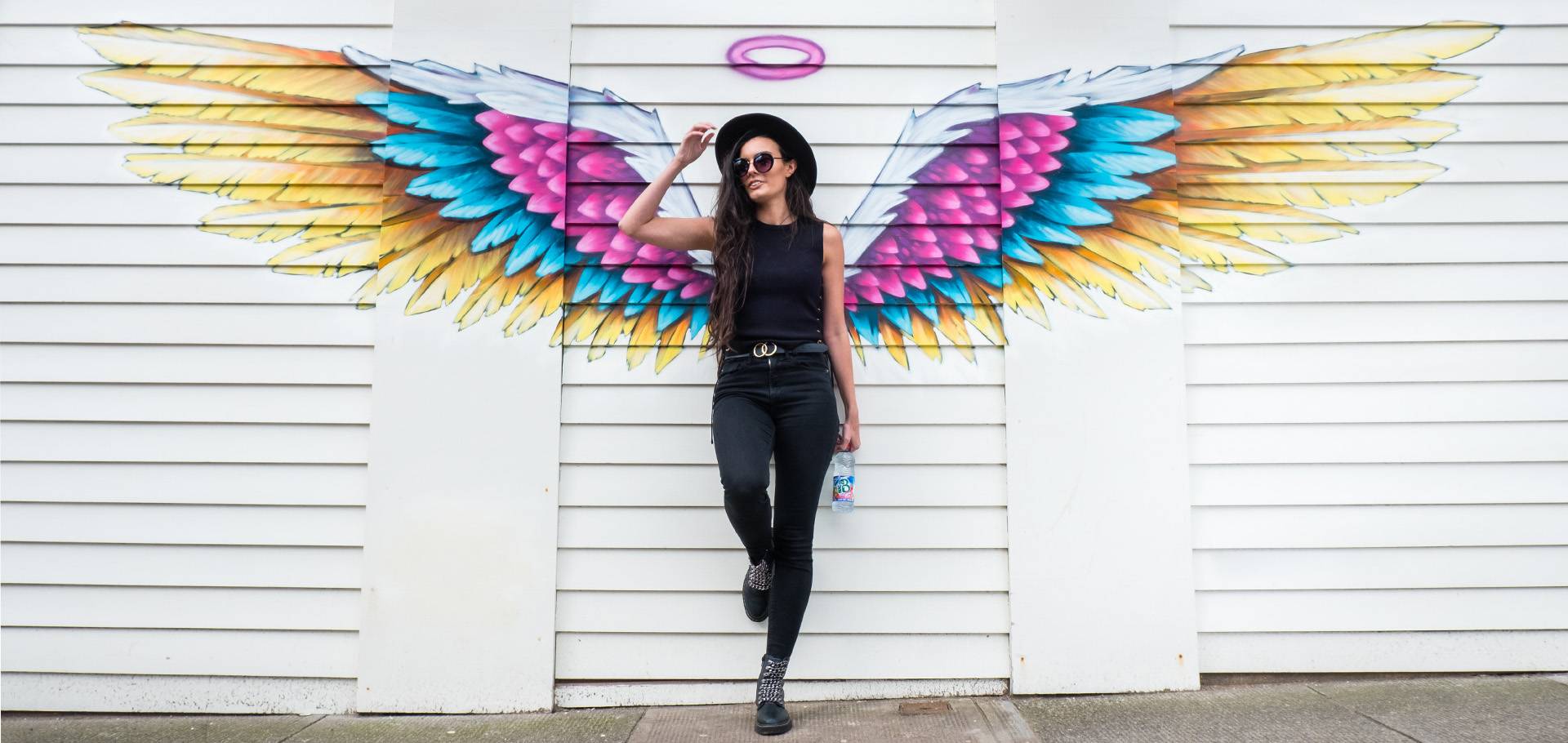 Mac-b photoshoot, a woman holding a bottle in front of a wing graphic on a wall