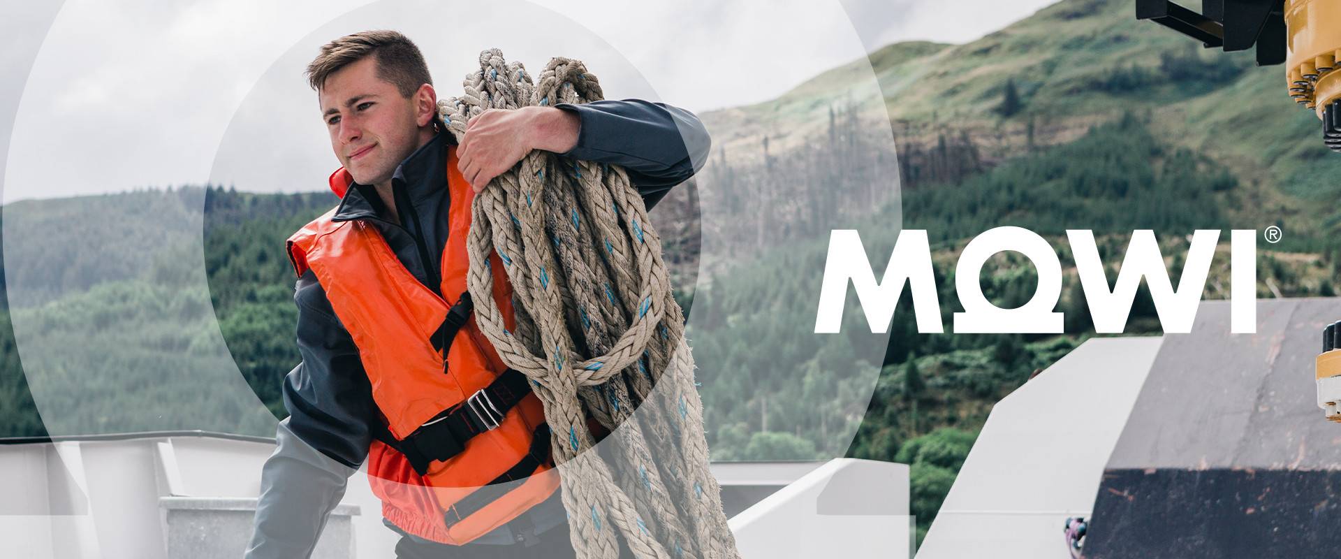 A man wearing a Mowi lifejacket on a boat, hauling some rope on his shoulder and the Scottish Highlands behind